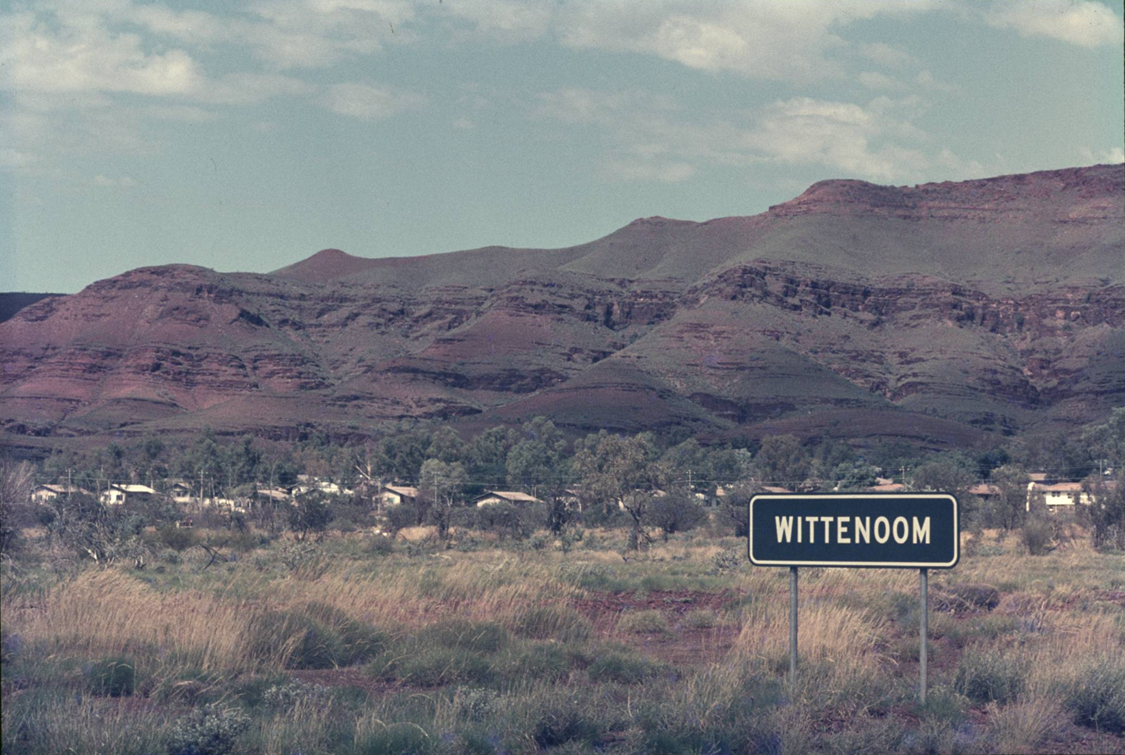 White text on a green sign. Sign says WITTENOOM. Houses, scrub, and hills visible in background.