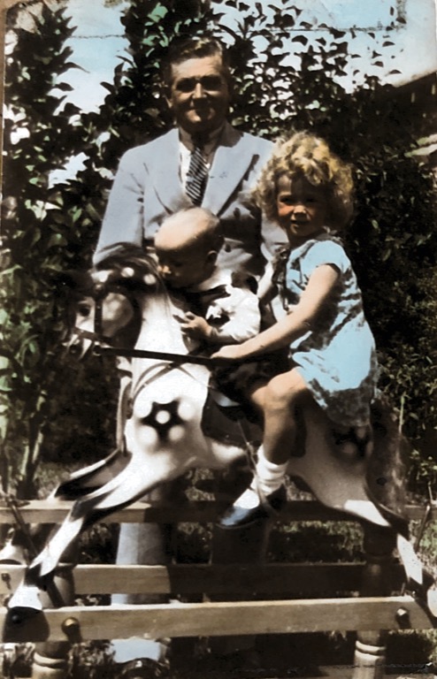 colourised photo of girl and bay on toy horse. A man is standing behind them.